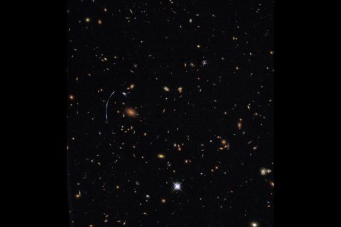 The galaxy cluster SDSS J1110+6459 is located about 6 billion light-years from Earth and contains hundreds of galaxies. At left, a distinctive blue arc is actually composed of three separate images of a more distant background galaxy called SGAS J111020.0+645950.8. The background galaxy has been magnified, distorted, and multiply imaged by the gravity of the galaxy cluster in a process known as gravitational lensing. (NASA, ESA, and T. Johnson (University of Michigan))