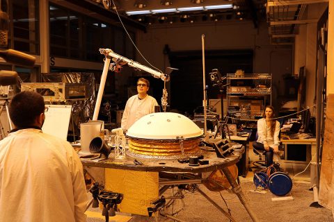 NASA's InSight mission tests an engineering version of the spacecraft's robotic arm in a Mars-like environment at NASA's Jet Propulsion Laboratory. The five-fingered grapple on the end of the robotic arm is lifting up the Wind and Thermal Shield, a protective covering for InSight's seismometer. The test is being conducted under reddish "Mars lighting" to simulate activities on the Red Planet. (NASA/JPL-Caltech)