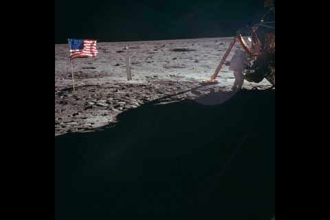 One of the few still photographs of Armstrong on the lunar surface, packing the bulk sample at the MESA; the American flag and the Solar Wind Collection experiment can be seen in the left of the photograph. (NASA)