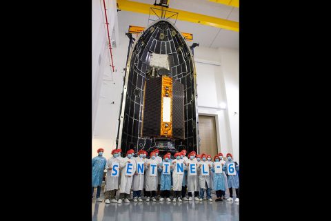 Technicians and engineers working on the Sentinel-6 Michael Freilich satellite pose in front of the spacecraft in its protective nosecone, or payload fairing, at Vandenberg Air Force Base in California. Once closed, the fairing will sit atop a SpaceX Falcon 9 rocket, launching in late November 2020. (NASA/Randy Beaudoin)