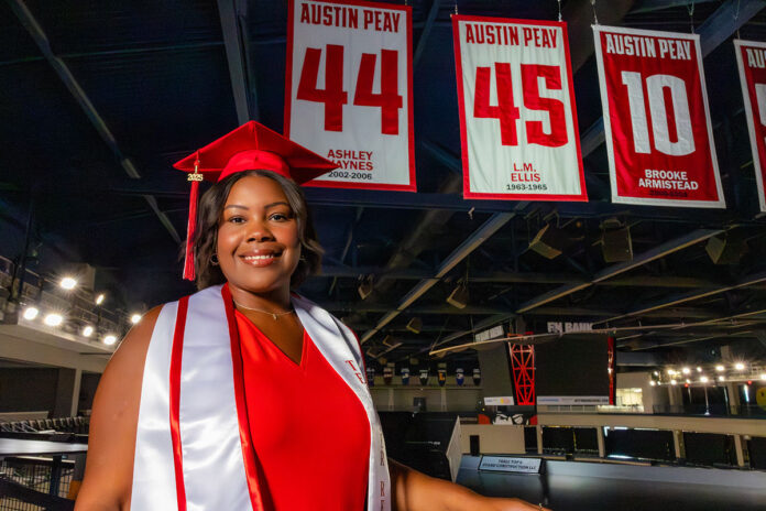 Jzsade Davenport, a Clarksville-Montgomery County School System teacher apprentice graduating from Austin Peay State University with her bachelor’s degree in elementary education this Friday, poses in front of the L.M. Ellis banner at F&M Bank Arena. Ellis’s jersey number was retired in 2022, to honor his place as the first Black player in APSU basketball history. Davenport is his granddaughter. (Robyn White, APSU)
